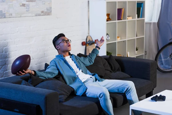 Emotionaler junger asiatischer Mann hält Rugby-Ball und schaut auf, während er auf der Couch sitzt und sich zu Hause ein Sportspiel anschaut — Stockfoto