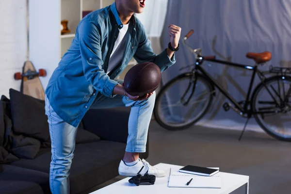 Schnappschuss eines aufgeregten jungen Mannes, der einen Rugby-Ball in der Hand hält und sich zu Hause ein Sportspiel anschaut — Stockfoto