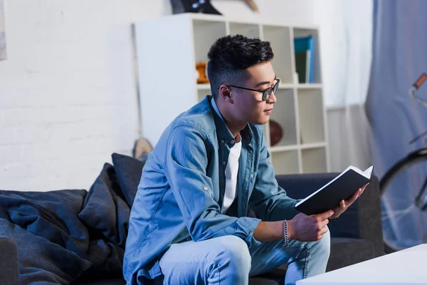 Joven asiático hombre en gafas sentado en sofá y lectura libro - foto de stock