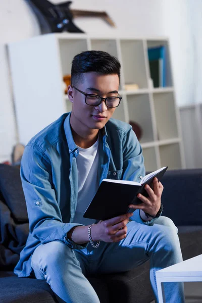 Guapo sonriente joven asiático hombre en gafas sentado en sofá y lectura libro - foto de stock