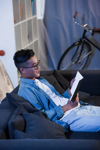 High angle view of smiling young man in eyeglasses sitting on couch and reading book — Stock Photo