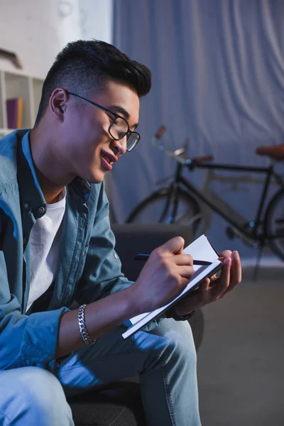 Усміхнений молодий азіатський чоловік в окулярах пише в блокноті вдома — стокове фото