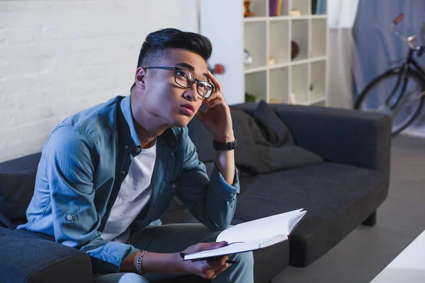 Pensativo jovem asiático homem em óculos segurando notebook e olhando para longe em casa — Fotografia de Stock
