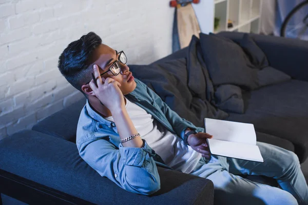 High angle view of pensive young asian man in eyeglasses holding notebook and looking away at home — Stock Photo