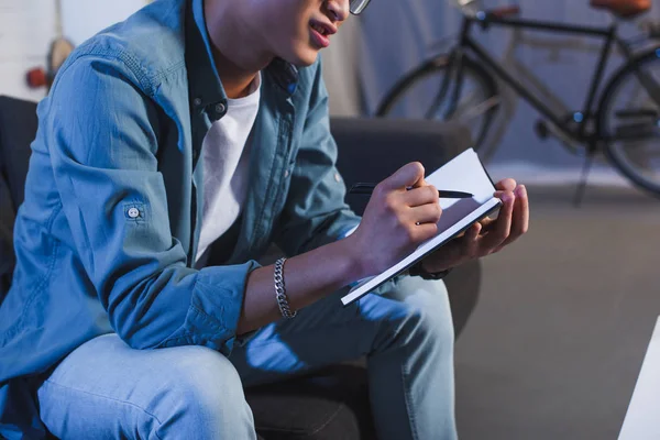 Обрізаний знімок молодого чоловіка, що сидить на дивані і пише в блокноті — стокове фото