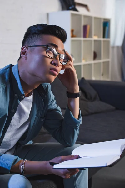 Pensive young asian man in eyeglasses holding notebook and looking away at home — Stock Photo