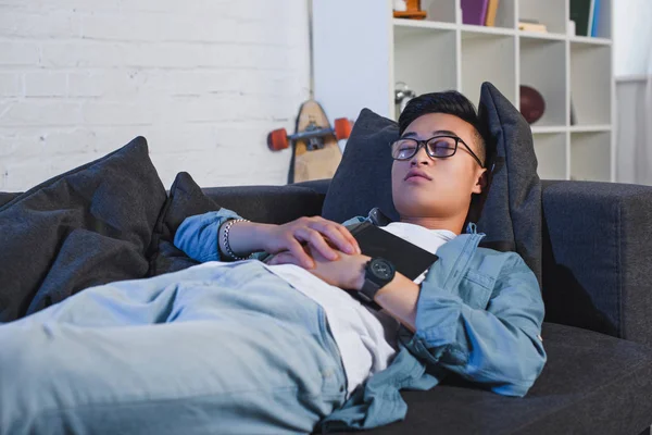 Young asian man in eyeglasses holding book and sleeping on couch — Stock Photo