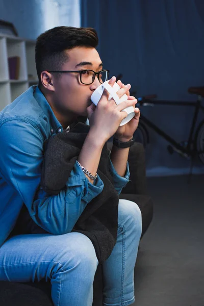 Guapo joven asiático hombre en gafas sentado en sofá y beber de taza - foto de stock