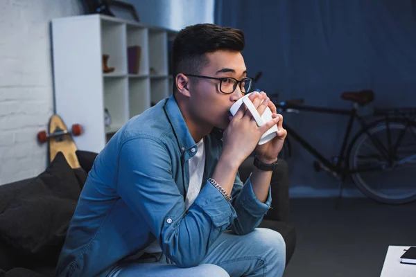 Junger asiatischer Mann in Brille sitzt auf Couch und trinkt aus Tasse — Stockfoto