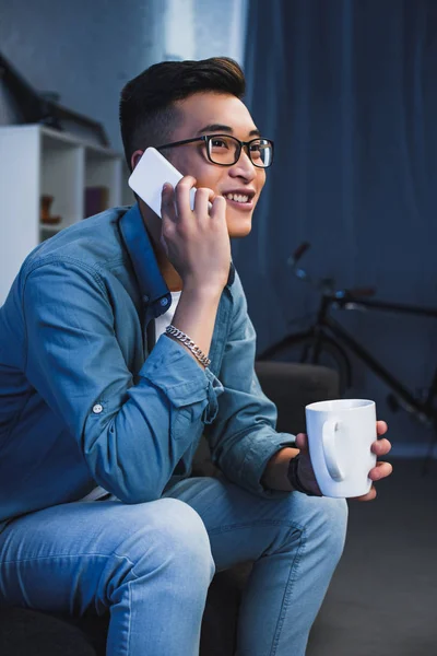 Smiling young asian man in eyeglasses holding mug and talking by smartphone — Stock Photo