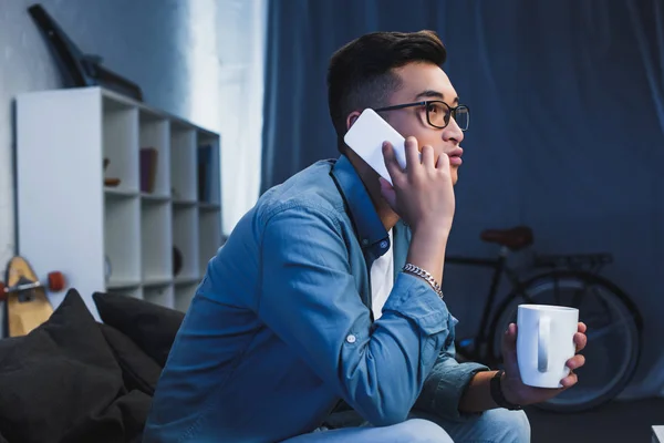 Vista lateral de joven asiático hombre en gafas holdign taza y hablando por teléfono inteligente - foto de stock