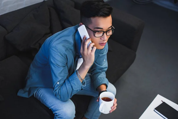 High angle view of young asian man in eyeglasses holdign cup of tea and talking by smartphone — Stock Photo