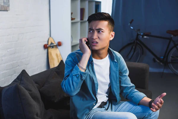 Upset young man sitting on couch and talking by smartphone at home — Stock Photo