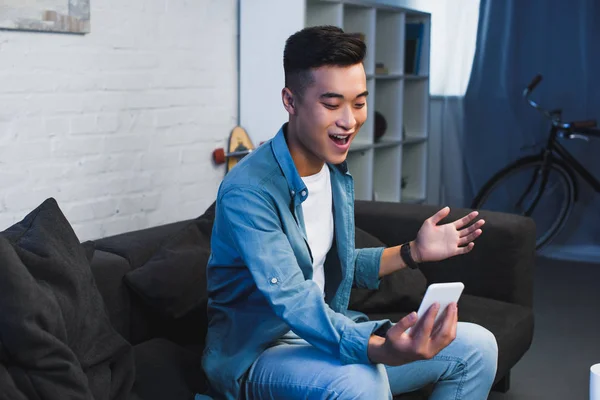 Sonriente joven asiático hombre sentado en sofá y el uso de teléfono inteligente en casa - foto de stock