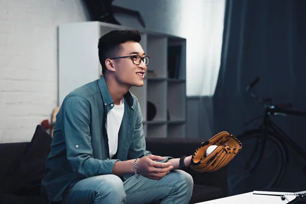 Lächelnder junger asiatischer Mann mit Baseballhandschuh und Ball sitzt auf der Couch und schaut sich zu Hause ein Sportspiel an — Stockfoto