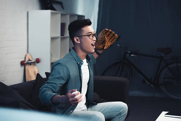 Emotionaler junger asiatischer Mann mit Baseballhandschuh und Ball sitzt auf der Couch und schaut sich zu Hause ein Sportspiel an — Stockfoto