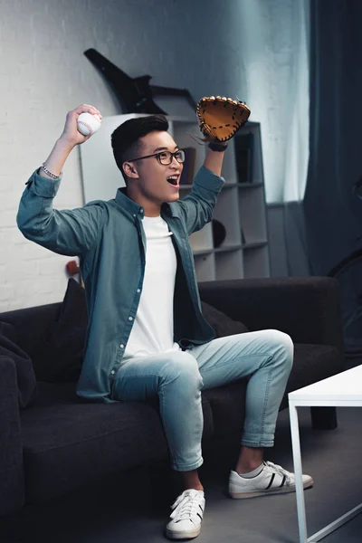 Eccitato giovane uomo con guanto da baseball e palla seduta sul divano e guardare la partita di sport a casa — Foto stock