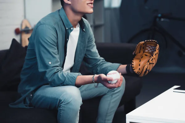 Recortado tiro de hombre joven con guante de béisbol y bola sentado en el sofá y viendo el deporte partido en casa - foto de stock