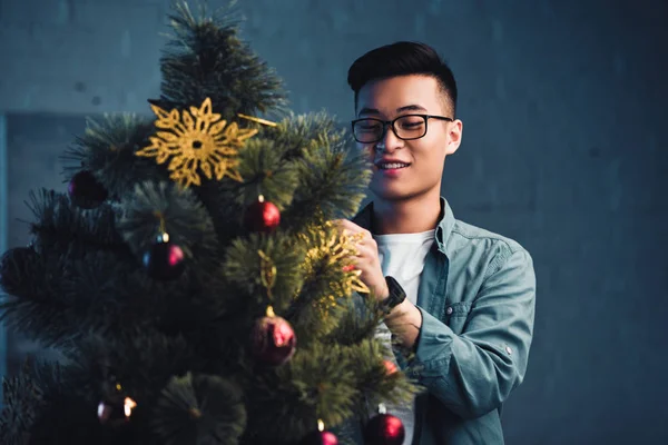 Smiling young asian man in eyeglasses decorating christmas tree at home — Stock Photo