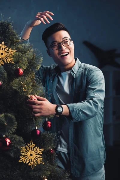Guapo joven asiático hombre riendo y decorando árbol de navidad - foto de stock