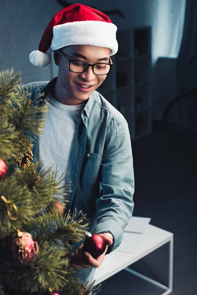 Handsome smiling young asian man in santa hat decorating christmas tree at home — Stock Photo