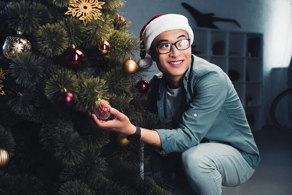 Guapo sonriente joven asiático hombre en santa hat decorando árbol de navidad y mirando hacia otro lado - foto de stock