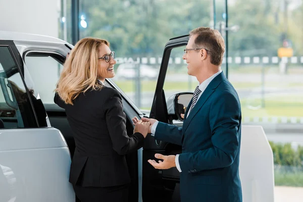 Smiling businessman helping businesswoman to sit into new car in showroom — Stock Photo