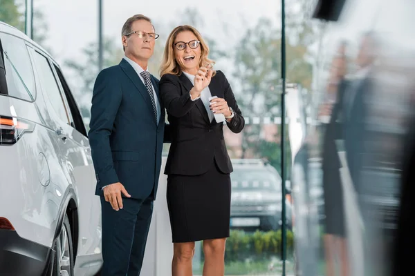 Femme d'affaires avec smartphone pointant du doigt tout en recherchant une nouvelle automobile avec un homme d'affaires dans le salon de concession — Photo de stock
