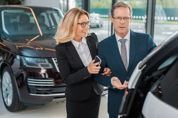 Homme d'affaires et femme d'affaires souriante avec smartphone choisir une nouvelle voiture dans le salon de concession — Photo de stock