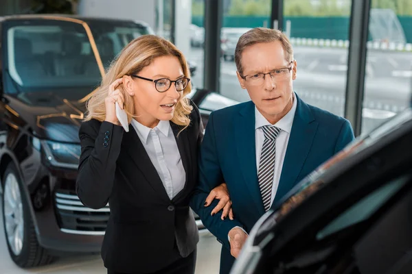 Businessman and businesswoman with smartphone choosing new car in dealership salon — Stock Photo