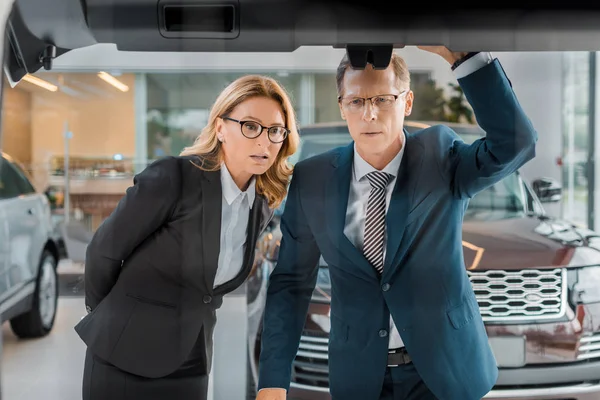 Pensive couple in formal wear looking at new car in dealership salon — Stock Photo