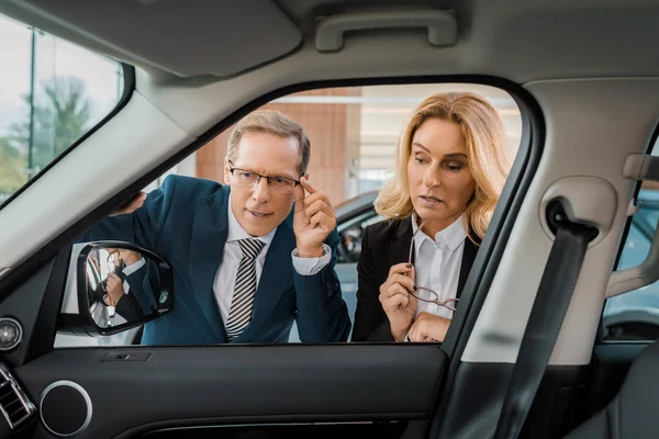 Hombre de negocios y mujer de negocios revisando coche nuevo en salón de concesionarios - foto de stock