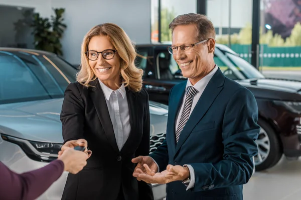 Partial view of female car dealer giving car key to smiling couple in formal wear in dealership salon — Stock Photo