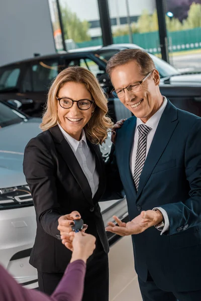 Partial view of female car dealer giving car key to smiling business couple in formal wear in dealership salon — Stock Photo