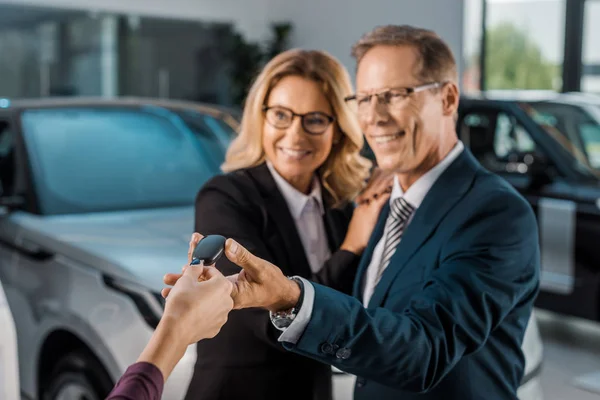 Vista parcial de concesionario de coche femenino dando la llave del coche a la pareja sonriente en el desgaste formal en salón de concesionarios - foto de stock