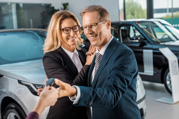 Partial view of female car dealer giving car key to smiling business couple in formal wear in dealership salon — Stock Photo