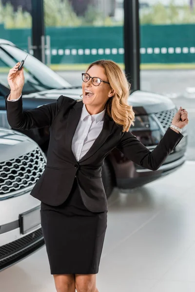 Mujer de negocios feliz en el desgaste formal que sostiene la llave del coche del coche nuevo en salón de concesionarios - foto de stock