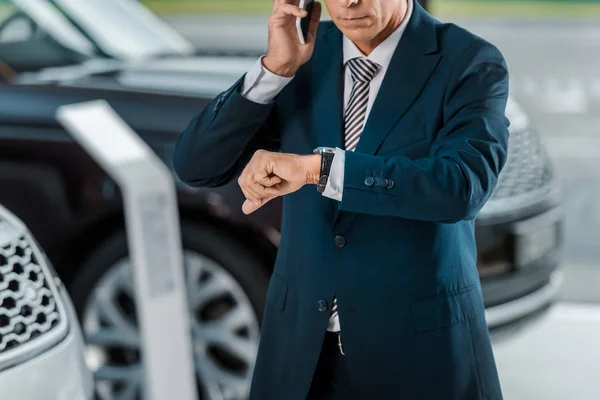 Cropped shot of adult businessman talking by phone at car dealership salon and looking at wrist watch — Stock Photo