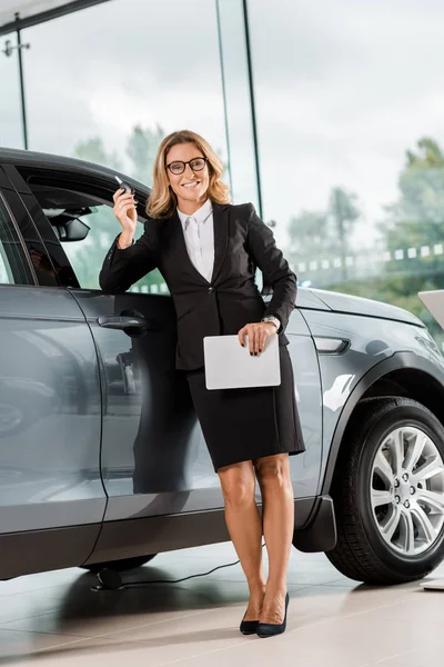 Sorridente rivenditore di auto femminile con tablet appoggiato alla macchina in showroom e guardando la fotocamera — Foto stock