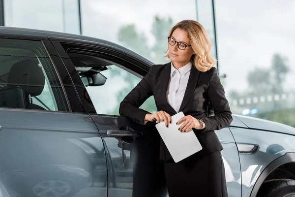 Beautiful female car dealer with tablet leaning on car in showroom — Stock Photo