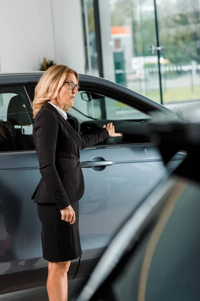 Attractive female car dealer pointing at car in showroom — Stock Photo