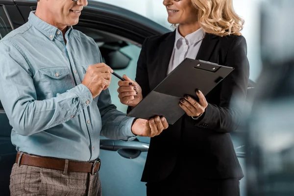 Обрезанный снимок взрослого клиента и продавщицы автомобилей с контрактом, стоящим в выставочном зале — стоковое фото