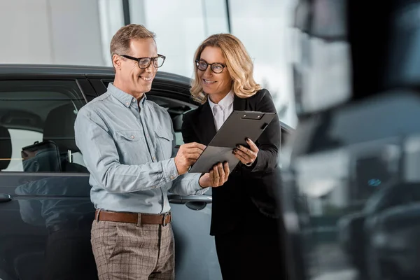 Hombre adulto y distribuidor de coche femenino con contrato de pie en la sala de exposición - foto de stock