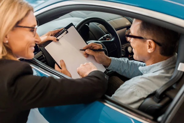 Happy adult man signing contract with female car dealer while sitting in car — Stock Photo
