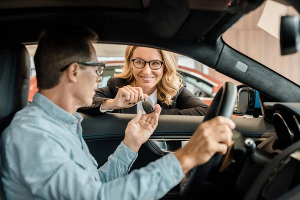Adult man taking car key from female car dealer at showroom while sitting inside car — Stock Photo