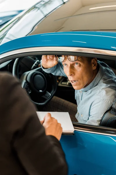 Обрезанный снимок женщины-автодилера, показывающей контракт автосалона с шокированным взрослым мужчиной в выставочном зале — стоковое фото