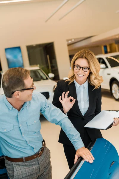 Homme adulte vérifiant voiture de sport de luxe avec concessionnaire de voiture femelle au showroom alors qu'elle montre signe correct — Photo de stock