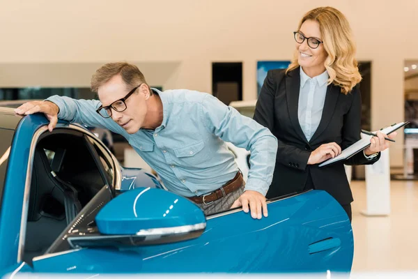 Hombre adulto que comprueba coche deportivo de lujo con distribuidor de coche femenino en sala de exposición - foto de stock