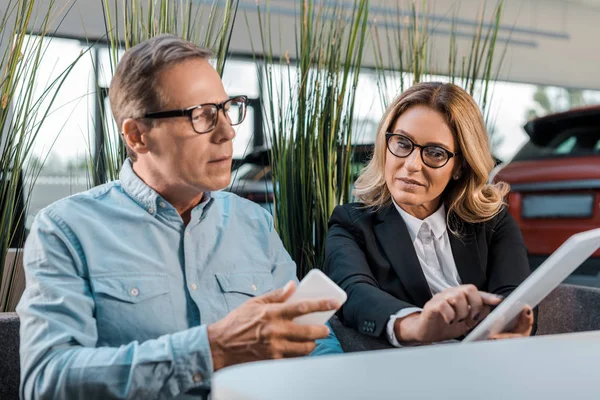 Homme adulte et femme concessionnaire de voitures avec téléphone et tablette assis au showroom — Photo de stock