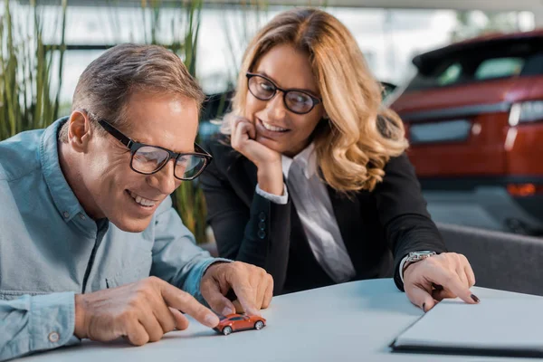 Happy adult man and female car dealer playing with toy car in showroom — Stock Photo